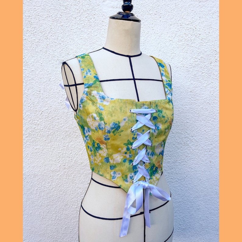 May include: A yellow and blue floral print corset top with white satin ribbon lacing. The corset top has a square neckline and a fitted bodice.