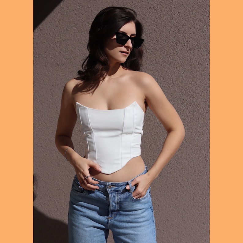 May include: A white strapless corset top with a pointed hemline. The top is made of a smooth, silky fabric and has a fitted silhouette. The model is wearing the top with blue jeans.
