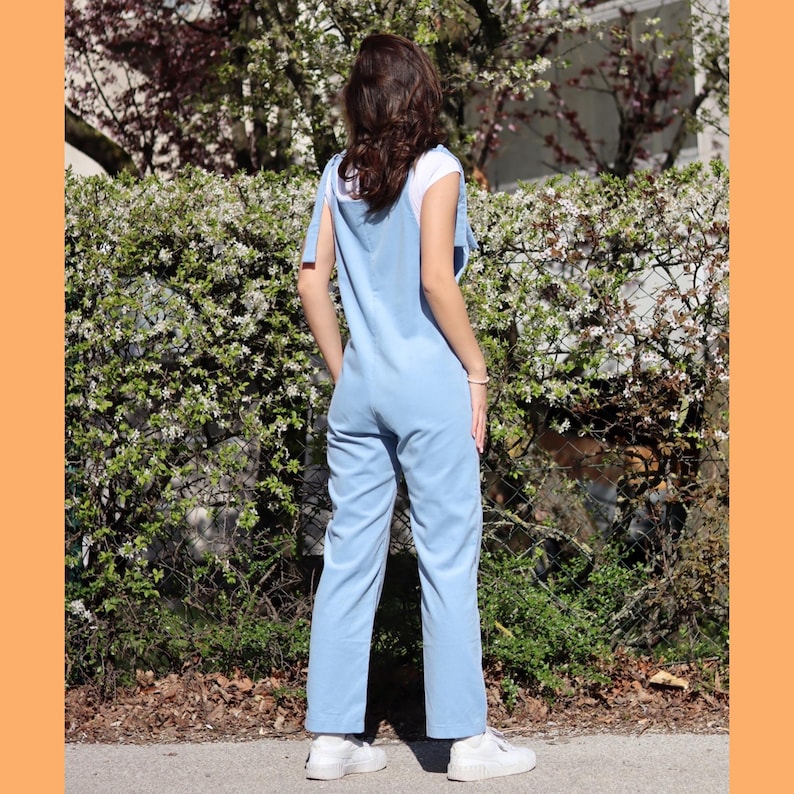 May include: A light blue overall jumpsuit with thin straps and a button closure. The straps are tied into a bow at the shoulders. The jumpsuit has a relaxed fit and straight legs.