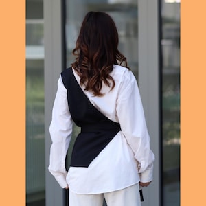 May include: A black vest with a diagonal design and a belt, worn over a white button-down shirt.