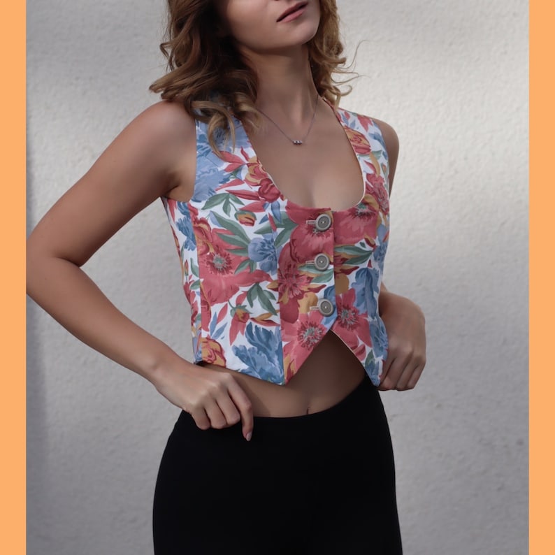 May include: A cropped floral print vest with a pointed hem and button closure. The vest is white with pink, blue, and yellow flowers.
