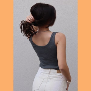 May include: A gray tank top with a scoop neck is worn with white jeans. The person is holding their dark brown hair up. A silver necklace and bracelet are visible. The background is a plain white wall with an orange border.