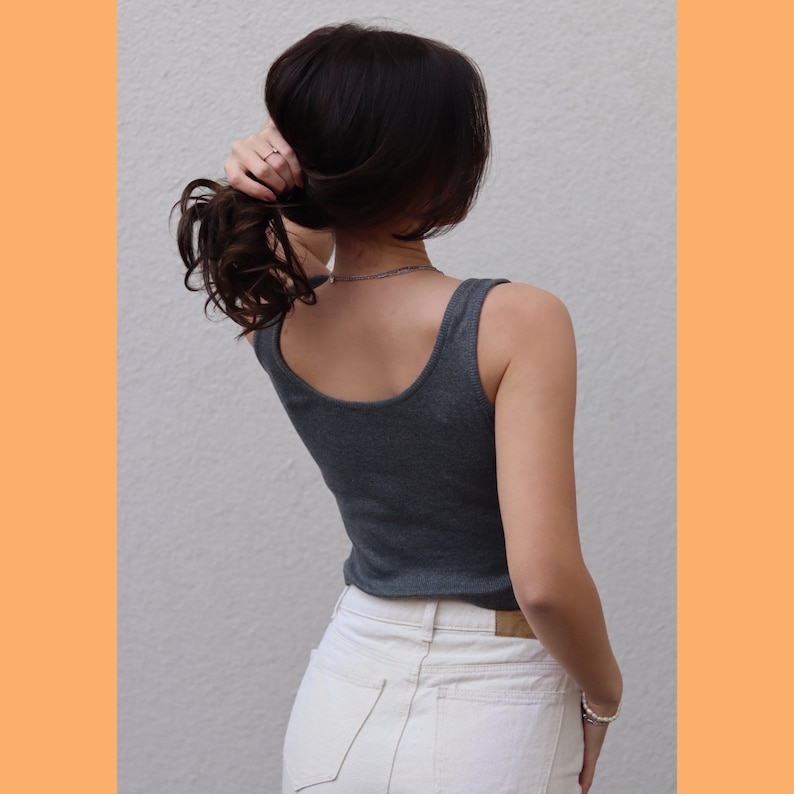 May include: A gray tank top with a scoop neck is worn with white jeans. The person is holding their dark brown hair up. A silver necklace and bracelet are visible. The background is a plain white wall with an orange border.