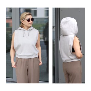 Puede incluir: Una sudadera con capucha sin mangas con capucha y cord&oacute;n. La sudadera es de color gris claro y se lleva sobre unos pantalones marrones.