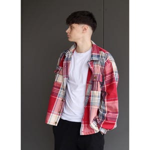 Pu&ograve; includere: Una camicia a quadri rossa, bianca e blu sopra una maglietta bianca. La camicia ha un'abbottonatura frontale e due tasche sul petto. La persona indossa pantaloni neri.