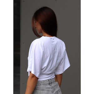 May include: White short-sleeved top with a wrap-around design. The top is paired with gray denim shorts. The model's back is facing the camera, with long brown hair.