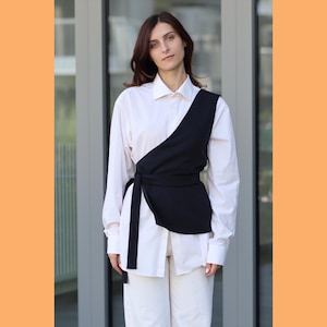 May include: A white button-down shirt with a black wrap top over it. The wrap top has a tie at the waist and a long, flowing sash.