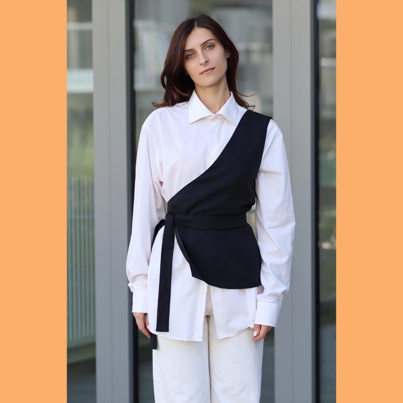 May include: A white button-down shirt with a black wrap top over it. The wrap top has a tie at the waist and a long, flowing sash.