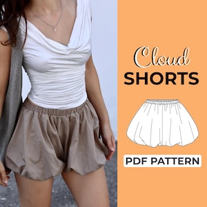 May include: A person wearing a white cowl neck top and brown shorts. The shorts are gathered at the waist and have a voluminous, rounded shape. The image also includes a graphic with the text "Cloud SHORTS" and "PDF PATTERN" on an orange background.