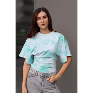 May include: A tie-dye t-shirt in shades of light blue and white. The shirt has short sleeves and a fitted waist. The model is wearing gray jeans. The background is a solid gray wall.