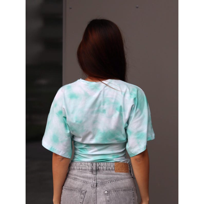 May include: A light blue and white tie-dye t-shirt with short sleeves. The shirt is paired with gray denim jeans. The model is facing away from the camera.