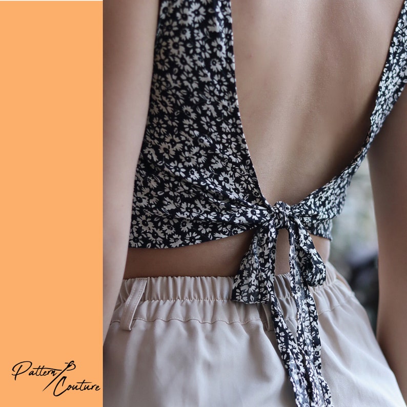 Op de afbeelding: Een zwart-wit gebloemd crop top met een lage rug en een strik sluiting. De top is aan de achterkant gestrikt en de uiteinden van de strik hangen tot aan de onderkant van de shorts. Het model draagt de top met beige shorts.