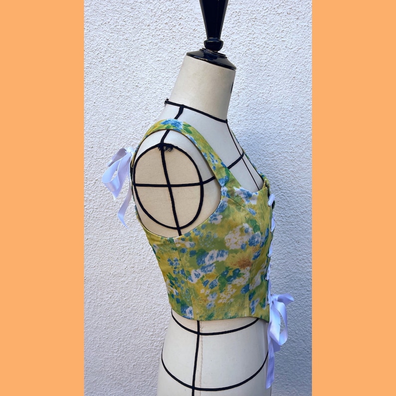 May include: A green and white floral print corset top with white ribbon ties. The top is on a white dress form.