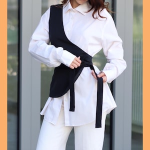 May include: A white button-down shirt with a black wrap top over it. The wrap top has a long tie that is tied at the waist.