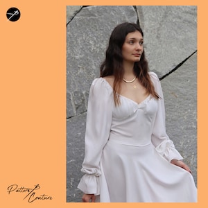 May include: A white dress with long sleeves and a sweetheart neckline. The dress has a ruffled trim around the neckline and cuffs. The model is wearing a gold necklace.