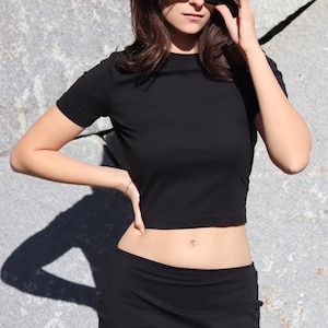 May include: A black cropped t-shirt with short sleeves and a black skirt. The model is wearing sunglasses.