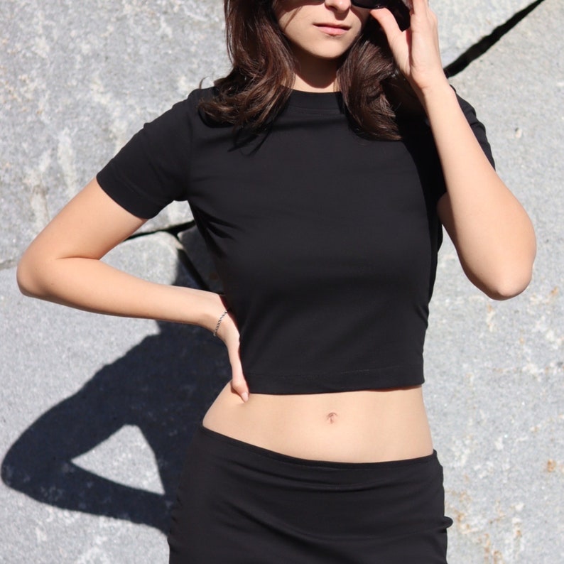 May include: A black cropped t-shirt with short sleeves and a black skirt. The model is wearing sunglasses.