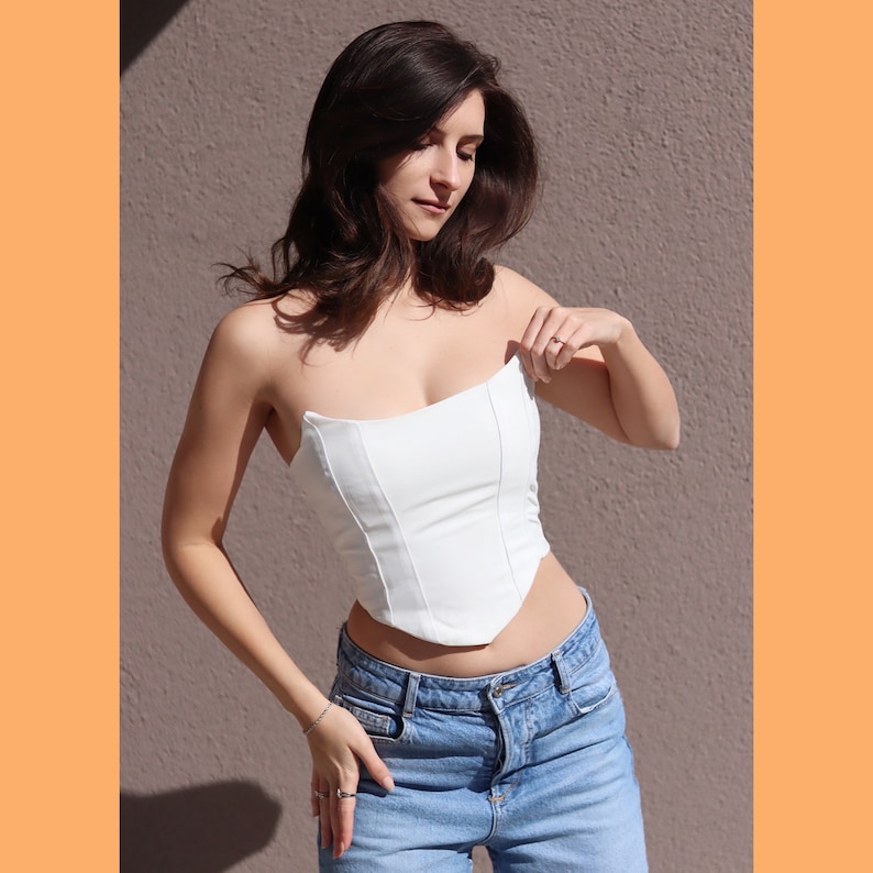 May include: A white strapless corset top with a pointed hemline. The top is made of a smooth, structured fabric and is worn with blue denim jeans.