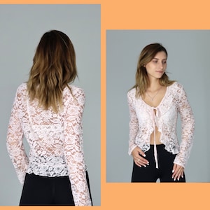 May include: A pink lace top with long sleeves and a tie at the neckline. The top is sheer and has a ruffled trim.