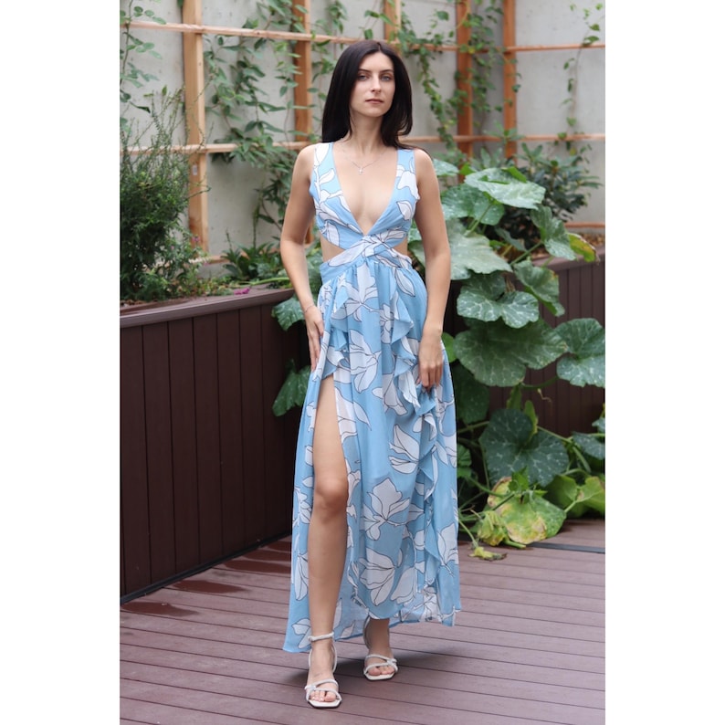 Op de afbeelding: Een lichtblauwe maxi-jurk met een witte bloemenprint. De jurk heeft een diep decollet&eacute;, uitsparingen in de taille en een hoge split aan de dij. De jurk is gemaakt van een lichtgewicht stof en heeft een zwierige silhouet.