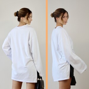 May include: A white long-sleeved shirt with a side slit. The shirt is loose-fitting and has a relaxed fit. The model is wearing the shirt with a black handbag.