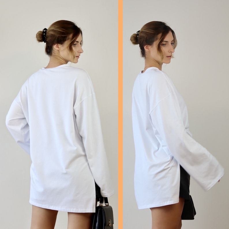 May include: A white long-sleeved shirt with a side slit. The shirt is loose-fitting and has a relaxed fit. The model is wearing the shirt with a black handbag.
