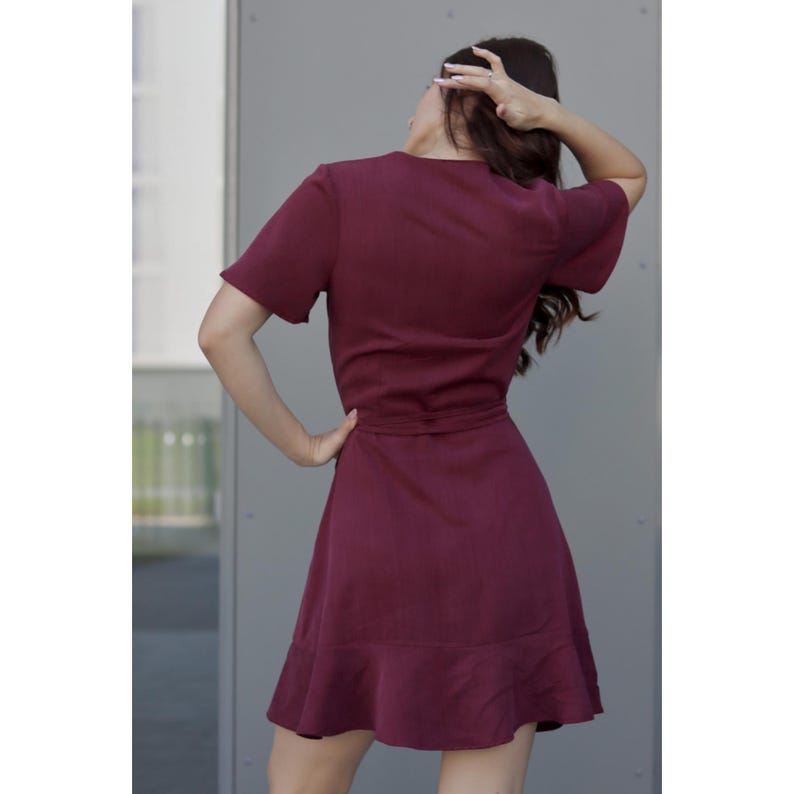 Peut inclure: Une robe portefeuille bordeaux &agrave; manches courtes et jupe &eacute;vas&eacute;e. La robe est dot&eacute;e d'une ceinture &agrave; la taille et d'un ourlet &agrave; volants. Le mod&egrave;le porte la robe, le dos tourn&eacute; &agrave; la cam&eacute;ra, la main dans les cheveux.
