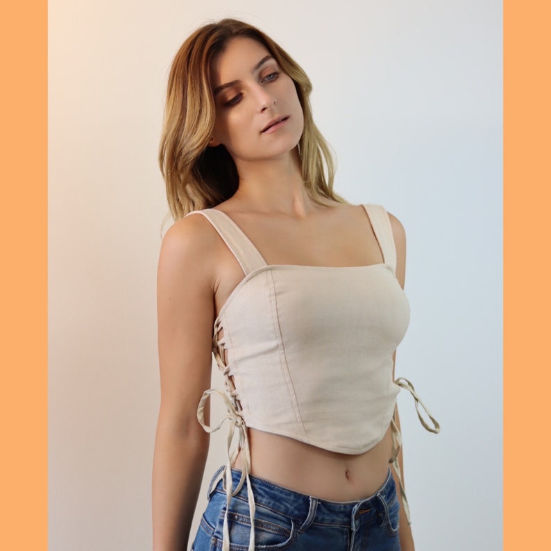 May include: A white linen crop top with a square neckline and lace-up detailing on the sides. The top is cropped above the waist and has a fitted silhouette.