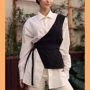 May include: A white button-down shirt with a black asymmetrical vest layered over it. The vest has a black belt that ties at the waist.