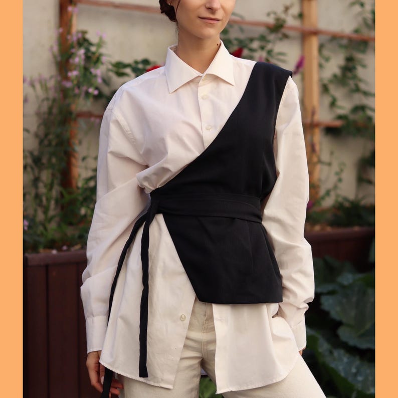 May include: A white button-down shirt with a black asymmetrical vest layered over it. The vest has a black belt that ties at the waist.