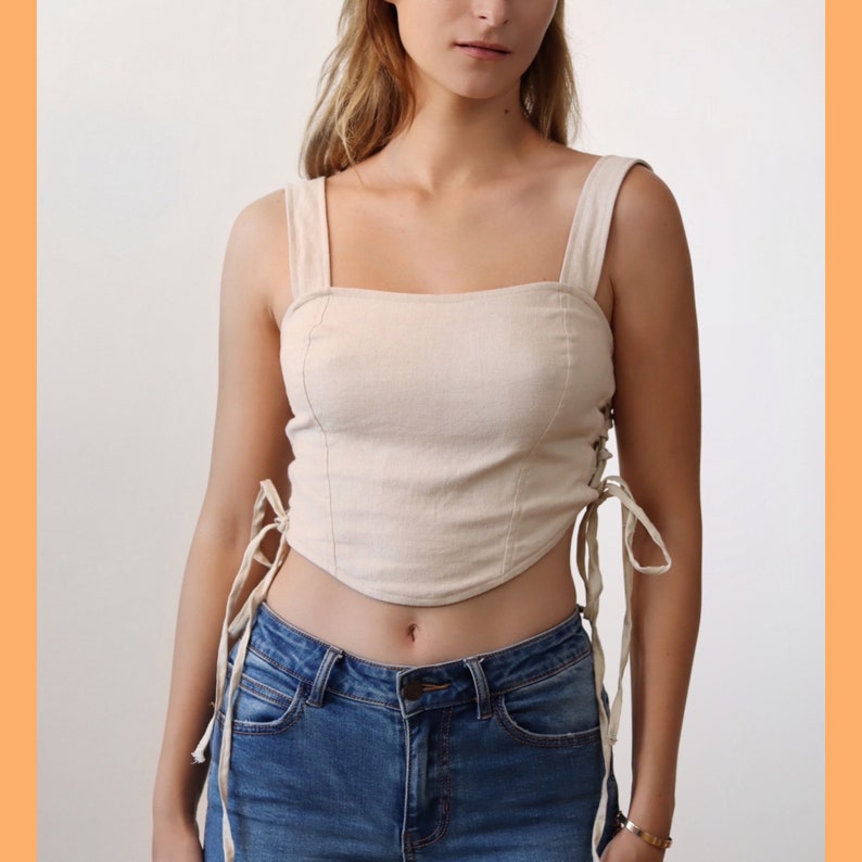 May include: A beige linen crop top with a square neckline and lace-up detailing on the sides. The top is tied at the waist and has a fitted silhouette.