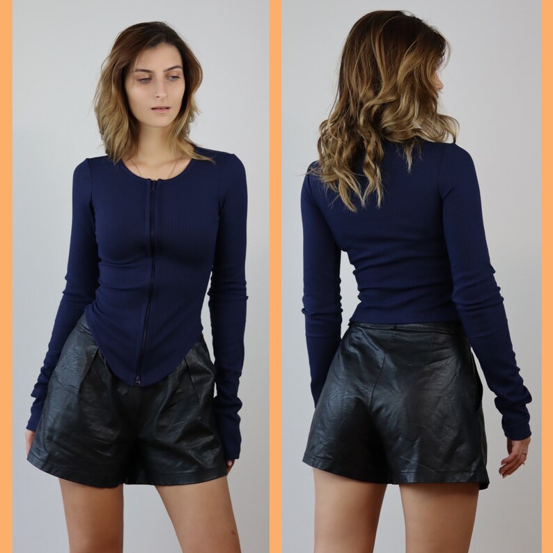 May include: A navy blue long-sleeved shirt with a zipper closure and a fitted silhouette. The shirt is paired with black leather shorts.