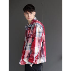 Pu&ograve; includere: Una camicia a quadri rossa, bianca e blu con una tasca sul petto. La camicia ha un'abbottonatura frontale e maniche lunghe. Il modello indossa pantaloni neri.