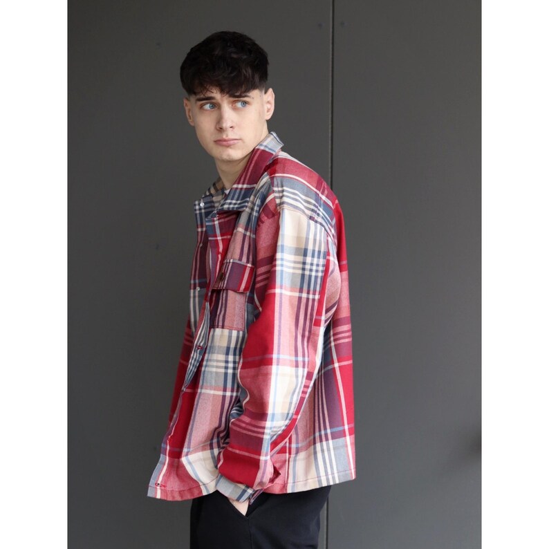 Pu&ograve; includere: Una camicia a quadri rossa, bianca e blu con una tasca sul petto. La camicia ha un'abbottonatura frontale e maniche lunghe. Il modello indossa pantaloni neri.