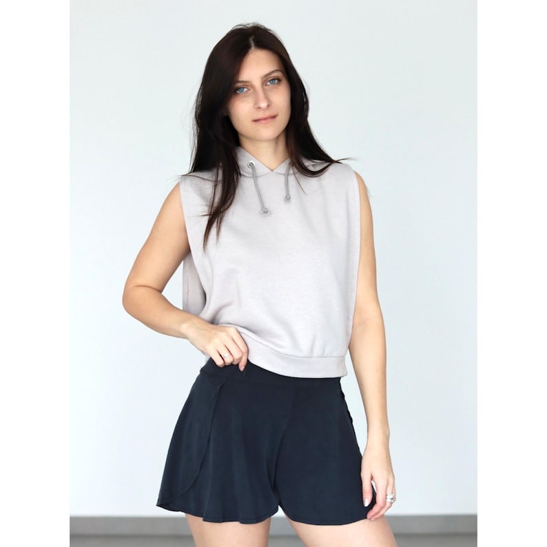 Puede incluir: Una sudadera sin mangas de color gris claro con capucha y cord&oacute;n de ajuste. La sudadera es corta y tiene un corte holgado. La modelo lleva pantalones cortos negros.