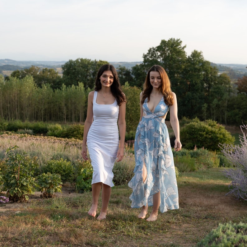Op de afbeelding: Twee vrouwen in zomerjurken lopen over een veld. De ene vrouw draagt een witte jurk en de andere een blauwe bloemenjurk.