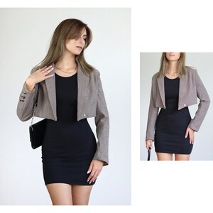 May include: A brown and black plaid cropped blazer with long sleeves and a single button closure. The blazer is worn over a black fitted dress.