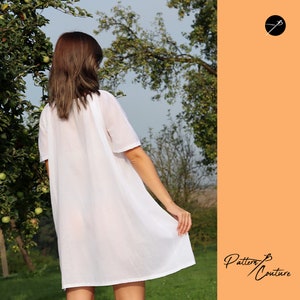 May include: A white, loose-fitting, short-sleeved dress with a round neckline. The dress is made of a lightweight fabric and has a flowing silhouette. The model is wearing the dress in a garden setting.