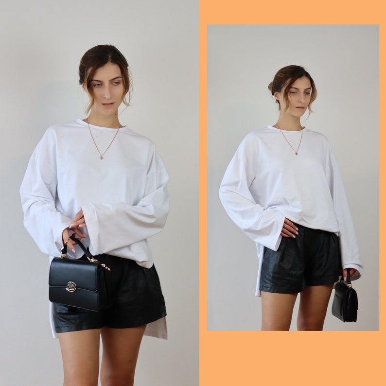 May include: A white long-sleeved shirt with a relaxed fit and a black leather shorts outfit. The person is wearing a gold chain necklace.