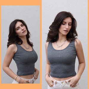 May include: A gray ribbed tank top with a scoop neck. The cropped top is paired with white pants and a silver necklace. The image is a studio shot with a neutral background.