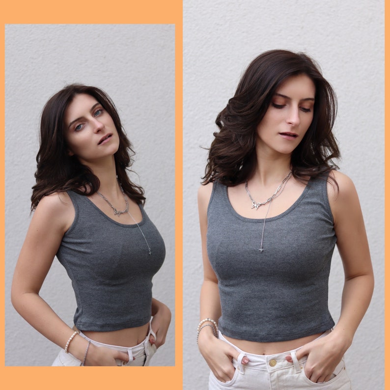 May include: A gray ribbed tank top with a scoop neck. The cropped top is paired with white pants and a silver necklace. The image is a studio shot with a neutral background.