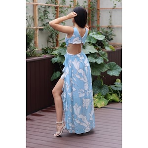 Op de afbeelding: Een lichtblauwe maxi-jurk met een witte bloemenprint en een open rug. De jurk heeft een geruite rok en een zijsplit.