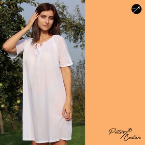 May include: A white, loose-fitting dress with a tie at the neckline. The dress has short sleeves and a flowing skirt. The model is standing in a garden setting.