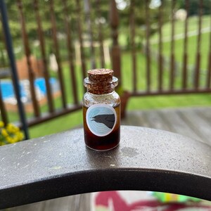 May include: A small glass bottle with a cork stopper and a black raven sticker. The bottle is filled with a dark brown liquid.