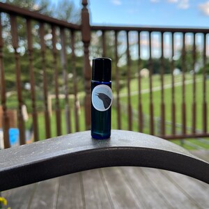 May include: A small, dark blue glass roller bottle with a black cap. The bottle has a label with a circular design featuring a black bird. The background is a blurred outdoor scene with a wooden deck and trees.
