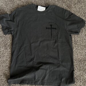May include: A dark grey t-shirt with a black graphic of a cross with nails and the text "Iron Sharpens Iron" around it.
