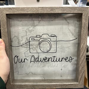 May include: A gray wooden frame with a glass front, displaying a vintage world map with a black line drawing of a camera and the text "Our Adventures".