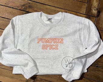 May include: A light gray sweatshirt with the words "PUMPKIN SPICE" embroidered in orange. The sweatshirt has a crew neck and long sleeves. A small circular logo with the letters "W D" is on the lower right side.