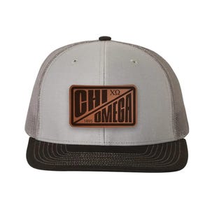 May include: A grey and black trucker hat featuring a rectangular brown leather patch on the front. The patch displays the text CHI OMEGA, the Greek letters Chi and Omega, and the year 1895, all engraved in a dark brown color.