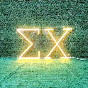May include: A neon sign displaying the Greek letters Sigma and Chi (ΣΧ) in a warm yellow glow. The letters are mounted on a clear backing, with a white power cord extending from the bottom right. The background is a textured green surface.